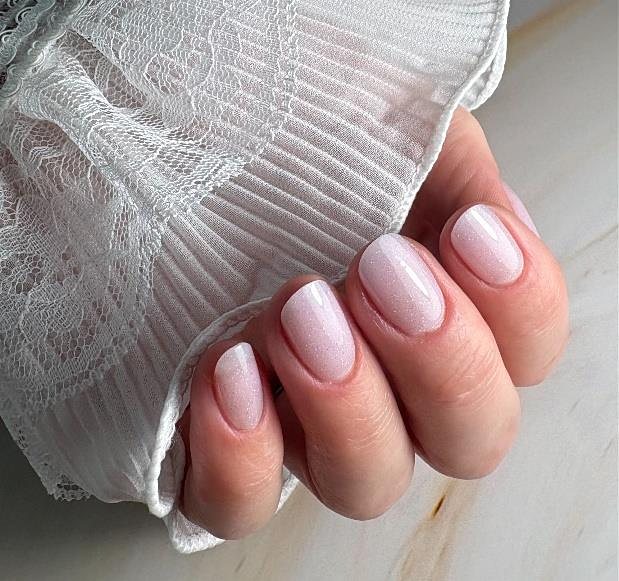 Zachtroze gelnagels op een hand met een witte kanten mouw.